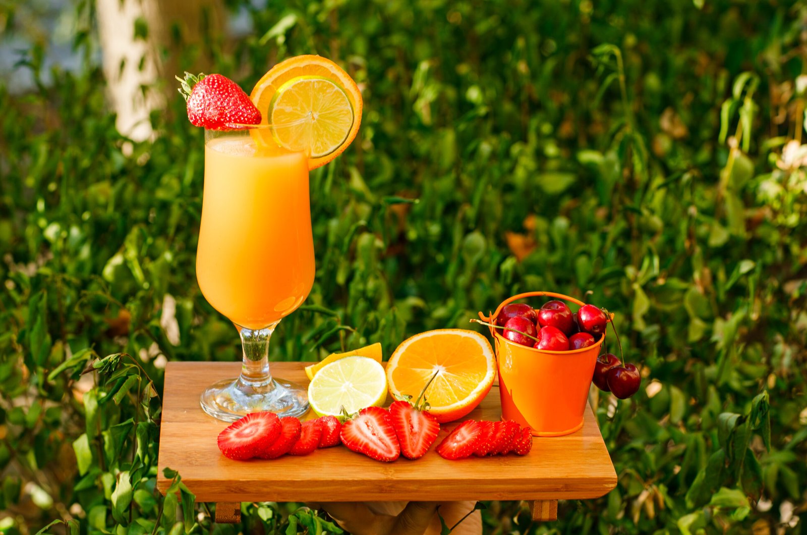 orange juice with citrus fruits, strawberry, cherry, cutting board in a goblet on meadow background, side view.